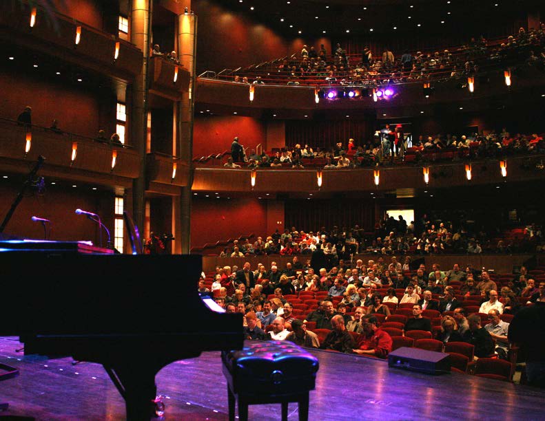 Audience seated in a multi-level theater, viewed from the stage beside a grand piano.
